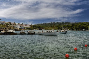 Menorca 'daki Cala Moli limanına demirlemiş teknelerin sakin manzarası, beyaz sahil evleri, yemyeşil alanlar ve arka planda dramatik bir gökyüzü..