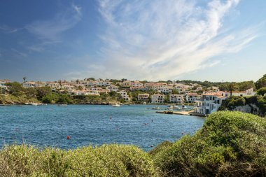 Menorca 'daki Cala Moli limanına demirlemiş teknelerin sakin manzarası, beyaz sahil evleri, yemyeşil alanlar ve arka planda dramatik bir gökyüzü..