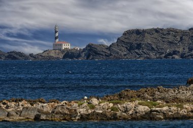 Striking view of Faro de Favaritx lighthouse perched on the rugged rocky coastline of Menorca, with a dramatic sky and deep blue sea. clipart