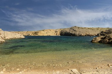Menorca 'da tenha bir koy olan Cala Pudenta' nın sakin manzarasında engebeli kayalık kayalıklarla çevrili turkuaz suları ve parçalı bulutlu bir gökyüzünün altındaki doğal manzaralar yer alıyor..