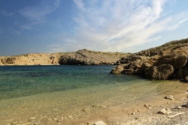 Menorca 'da tenha bir koy olan Cala Pudenta' nın sakin manzarasında engebeli kayalık kayalıklarla çevrili turkuaz suları ve parçalı bulutlu bir gökyüzünün altındaki doğal manzaralar yer alıyor..