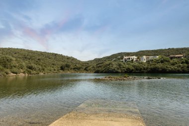 Menorca 'daki Cala Rata' da sakin sular, yemyeşil tepeler ve berrak mavi gökyüzü altında birkaç büyüleyici ev içeren huzurlu bir rıhtım sahnesi..