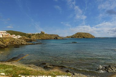 Menorca 'daki Cala Sa Mesquida' nın çarpıcı kıyı manzarası berrak mavi sular, kayalık kıyı şeridi ve parlak mavi bulutlar altında büyüleyici beyaz evler..