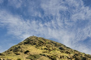 Menorca 'da Morro de Tramuntana tepesinde duran iki yürüyüşçü, engebeli yamacın geniş bir manzarası ve parlak mavi gökyüzü ince bulutlarla dolu..
