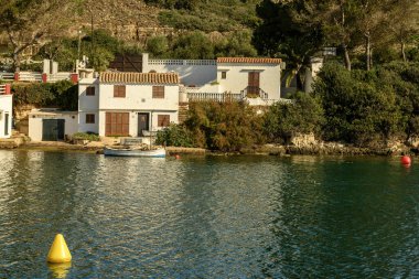 Menorca 'daki Cala Sant Esteve' de sakin bir limanda demirlemiş tekneler, yemyeşil alanlar ve bulutlarla dolu canlı bir gökyüzü altında büyüleyici evler yer alıyor..