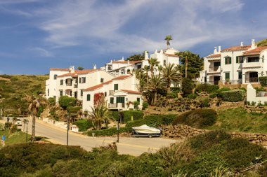 Menorca 'daki Cala Tirant plajının güzel manzarası, berrak mavi sular, kumlu kıyılar ve canlı mavi gökyüzünün altındaki yeşil tepelere kurulmuş beyaz evler..