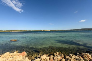 Menorca 'daki Cala de Fornells' in göz kamaştırıcı manzarası, kristal berrak turkuaz suları, kayalık kıyı şeridi ve parlak mavi bir gökyüzünün altında ufukta bir yelkenli..