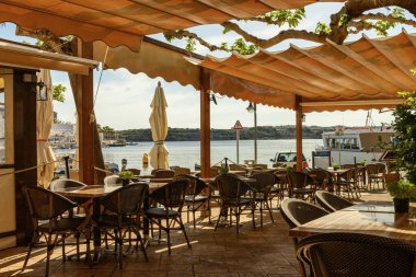 Menorca 'daki Dips Limanı' nda bir sahil restoranının açık hava oturma alanı. Limanı ve tekneleri gören bir gölgelik altında masa ve sandalyeler. Güneşli bir gün ve huzurlu bir ortam..