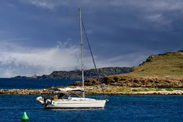 stock image A sailboat anchored in the clear blue waters near Faro de Favaritx lighthouse, Menorca. The rugged coastline and dramatic sky provide a stunning backdrop.