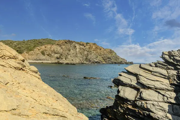 stock image Stunning coastal landscape of Cala Sa Mesquida in Menorca, featuring clear blue waters, rocky shoreline, and charming white houses under a vibrant blue sky with wispy clouds.