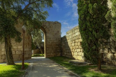 İspanya 'nın Merida kentindeki antik Roma duvarları, uzun selvi ağaçları ve açık mavi gökyüzünün altında bir sokak lambası ile çevrili. Tarihi taş yapılar şehrin zengin kültürel mirasını vurguluyor..