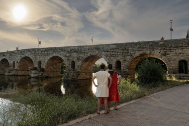 İspanya 'nın Merida kentindeki Guadiana Nehri üzerindeki antik Roma köprüsünün yanında Roma giysisi giymiş iki canlandırmacı. Sahne, gün batımında dramatik bir gökyüzü altında tarihi atmosferi yakalar..