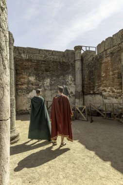 Two men dressed in Roman-style cloaks observe a statue at an ancient Roman archaeological site in Merida, Spain, surrounded by historic stone columns and walls. clipart