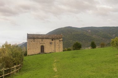 Kırsal bir tasarıma sahip taştan şapel, bulutlu bir gökyüzünün altında dağlık bir zemin ile yeşil bir alana kurulmuş..