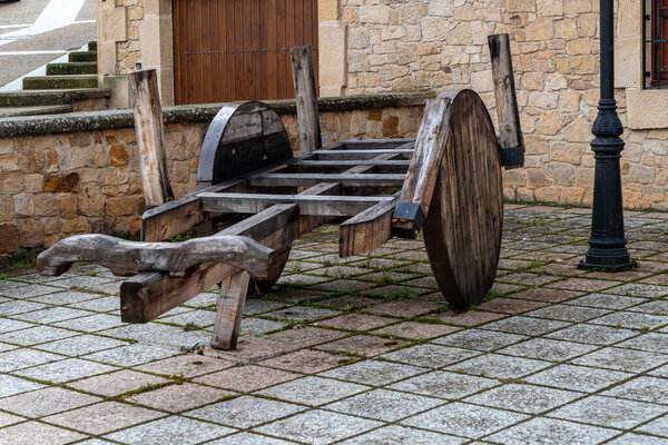 Традиционная деревянная картонка с большими колесами, установленная на мощеной улице в испанском городе Сория рядом с каменной стеной и светильником.