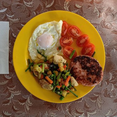 Kızarmış yumurta, ızgara burger köftesi, taze domates ve sarı tabakta sotelenmiş sebze içeren renkli bir yemek..