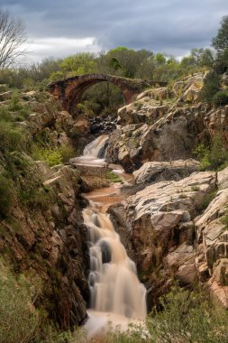 İspanya 'nın Vadollano kentindeki engebeli kayalar ve yemyeşil bitki örtüsüyle çevrili küçük bir şelalenin üzerinde zarif bir kemer açıklığına sahip tarihi bir taş köprü..
