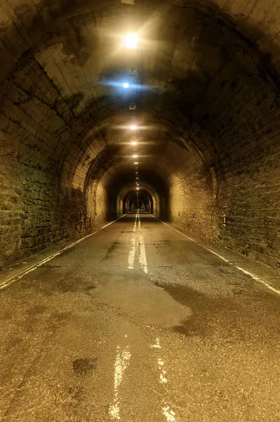 Taş duvarları ve merkezi beyaz çizgileri olan loş ışıklı bir yol tüneli derinlik ve perspektif hissi yaratıyor..