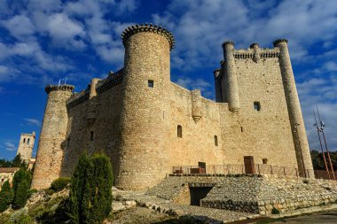 İkonik Castillo de Torija, sağlam taş duvarlar ve silindirik kuleler, canlı mavi bir gökyüzüne karşı dimdik ayakta duruyor ve yeşilliklerle çevrili..