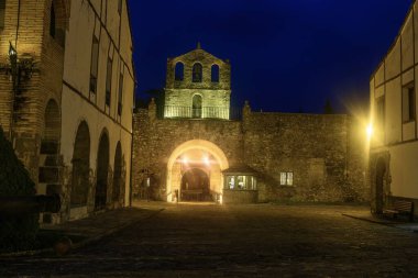 Bir çan kulesi ve kemerli girişi olan tarihi taş kale gece vakti koyu mavi gökyüzünün altında güzel aydınlatılıyor..