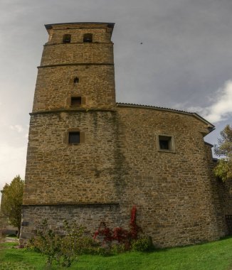 Bir saati ve kemerli girişi olan uzun taş çan kulesi, yeşilliklerle çevrili ve parlak mavi gökyüzüne karşı kurulmuş..