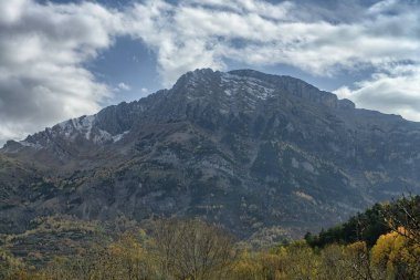 Yoğun sonbahar yaprakları ve çarpıcı bulutlu gökyüzü ile çevrili karla kaplı bir dağın dramatik manzarası..