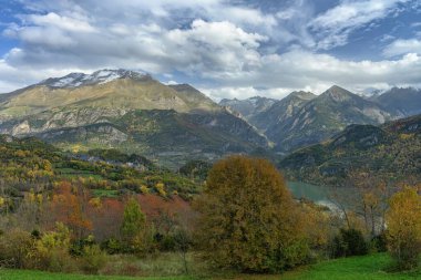 Yoğun sonbahar yaprakları ve çarpıcı bulutlu gökyüzü ile çevrili karla kaplı bir dağın dramatik manzarası..