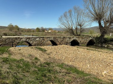 İspanya kırsalında küçük bir nehir boyunca uzanan iki kemerli eski Roma taş köprüsü.