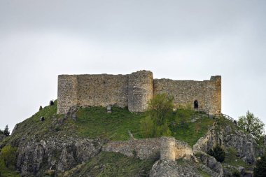 Yeşillik ve ahşap çitlerle çevrili kayalık bir tepedeki eski taş kale.