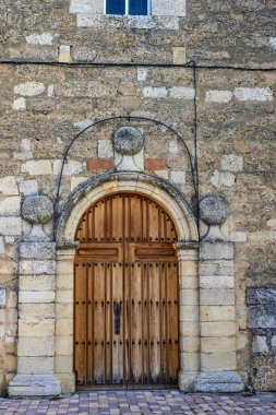Saat ve çan kulesi olan taş kilise, kemerli ahşap giriş ve köy meydanındaki budanlı ağaç.