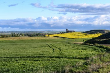 Palencia 'da dramatik bulutlu gökyüzü ile sarı bir çiçek tarlasının arkasında yükselen eski gotik kilise.