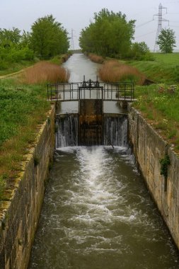 Yeşil bitki örtüsü ve tarım arazisi tarafından çerçevelenmiş, içinden akan suyla tarihi kanal kilidi