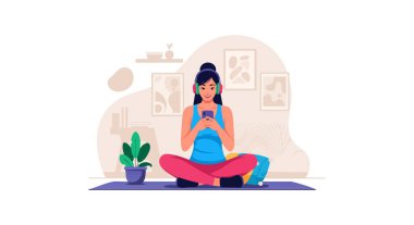 Yoga minderinde bacak bacak üstüne atmış, kulaklıklı akıllı telefona odaklanmış, bitkiler ve çerçeveli resimlerle çevrili, sakin ve huzurlu bir şekilde oturuyor..