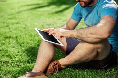 Genç hippi çocuk parkta oturuyor ve modern dijital tablet kullanıyor, erkek blogcu dışarıda tablet kullanıyor, sosyal ağ konsepti