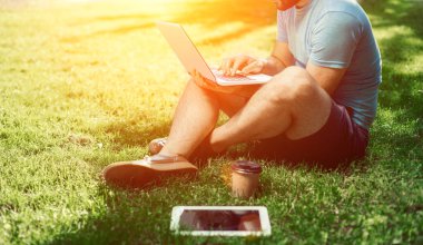 Genç blogcu çimlerde oturuyor ve dizüstü bilgisayarla çalışıyor. Uzayı kopyala Güneş parlaması