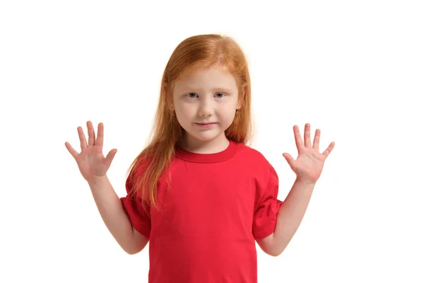女の子は腕を上げて微笑んだ 5歳の女の子の笑顔と手の近くに彼女の手に驚いている 白い背景に孤立した赤いTシャツの赤い髪の少女 — ストック写真