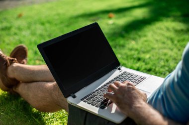 Cropped shot of man using laptop with blank screen while sitting on green grass. Freelance works in the open air in the park