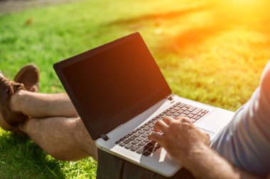 Cropped shot of man using laptop with blank screen while sitting on green grass. Freelance works in the open air in the park. Sun flare