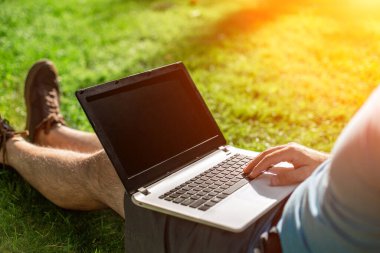 Cropped shot of man using laptop with blank screen while sitting on green grass. Freelance works in the open air in the park. Sun flare