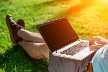 Cropped shot of man using laptop with blank screen while sitting on green grass. Freelance works in the open air in the park. Sun flare