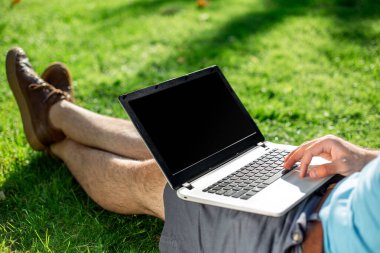 Cropped shot of man using laptop with blank screen while sitting on green grass. Freelance works in the open air in the park