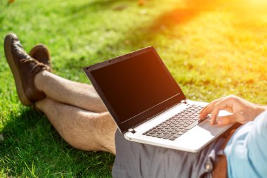 Cropped shot of man using laptop with blank screen while sitting on green grass. Freelance works in the open air in the park. Sun flare