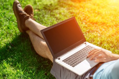 Cropped shot of man using laptop with blank screen while sitting on green grass. Freelance works in the open air in the park. Sun flare