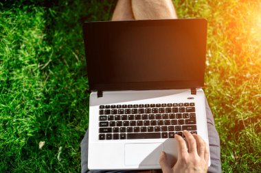 Cropped shot of man using laptop with blank screen while sitting on green grass. Freelance works in the open air in the park. Sun flare