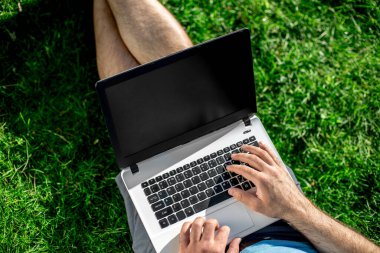 Cropped shot of man using laptop with blank screen while sitting on green grass. Freelance works in the open air in the park