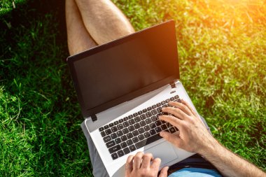 Cropped shot of man using laptop with blank screen while sitting on green grass. Freelance works in the open air in the park. Sun flare