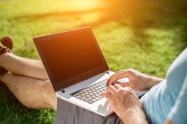 Cropped shot of man using laptop with blank screen while sitting on green grass. Freelance works in the open air in the park. Sun flare