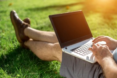 Cropped shot of man using laptop with blank screen while sitting on green grass. Freelance works in the open air in the park. Sun flare