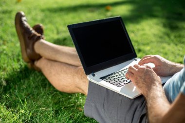 Cropped shot of man using laptop with blank screen while sitting on green grass. Freelance works in the open air in the park