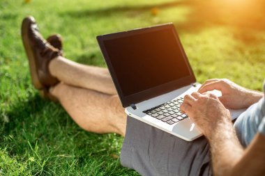 Cropped shot of man using laptop with blank screen while sitting on green grass. Freelance works in the open air in the park. Sun flare
